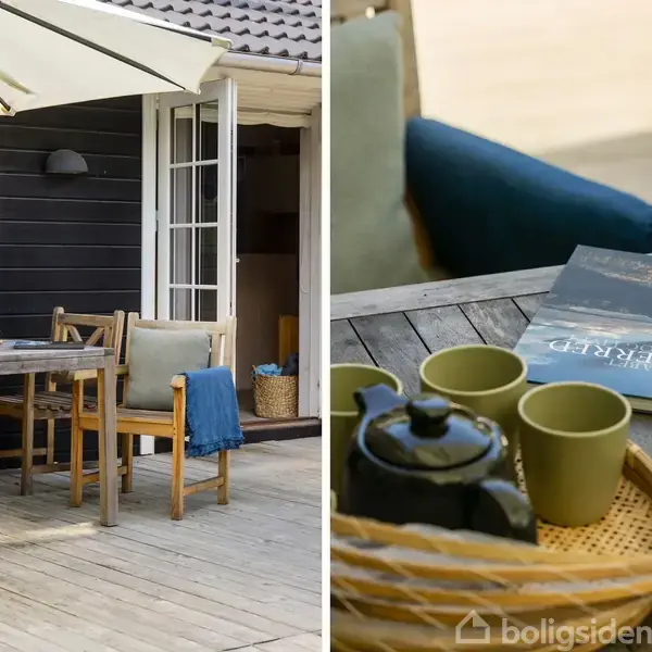 Udendørs træterrasse med spisebordssæt under stor parasol. Der er et åbent vindue mod et sort træhus. På bordet står tekande, krus og en bog med omslaget "DESTINATION SØSTRÆNDE," samt en blå pude.