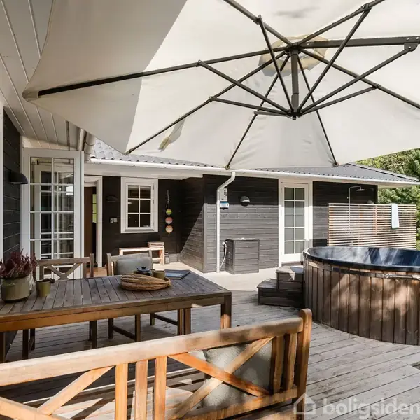 Træterrasse med rundt spabad står under stor parasol. Træbord med bænke foran vinduer fra huset i baggrunden. Grønne træer omkranser området, der skaber en afslappet udendørs oase.