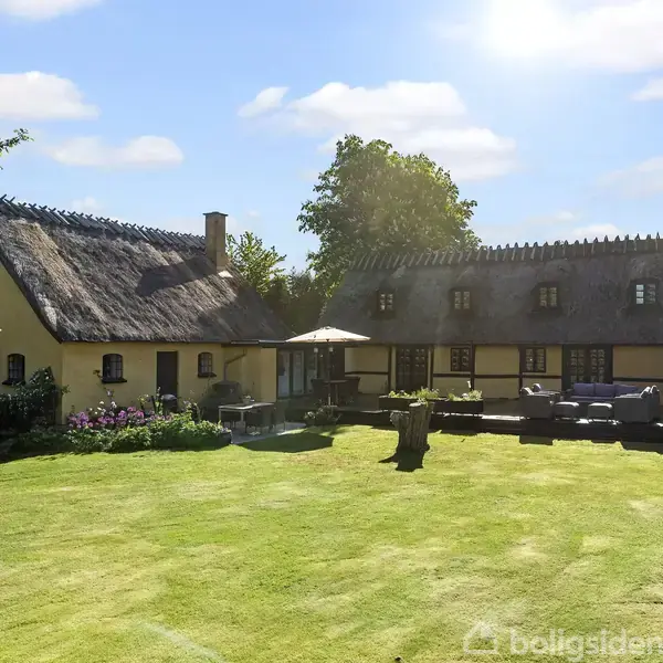 Et stråtækt hus står stille i en solbeskinnet have. Omkring huset ses grønne træer og en velplejet græsplæne med solstole og parasol.