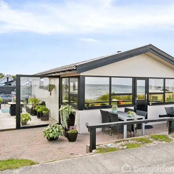 Et moderne sommerhus, omgivet af terrasse og blomsterkrukker, står med udsigt til havet. Store vinduer og udendørs siddepladser fremhæver en afslappet og naturskøn atmosfære.
