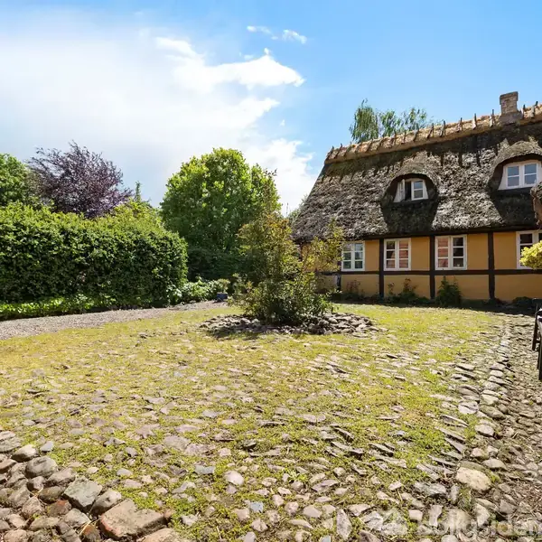 Bindingsværkshus står roligt med stråtækt tag og gule vægge. Grusbelagt gårdhave omkranser huset, omgivet af frodig vegetation og en blå himmel.