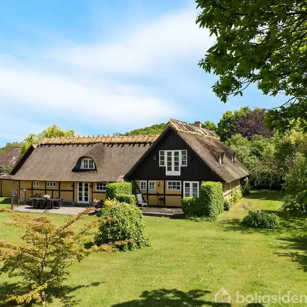 Stråtækt bindingsværkshus ligger midt i en grøn have med træer og buske, under en klar blå himmel. Huset har hvide vinduer og fremstår idyllisk og hyggeligt.