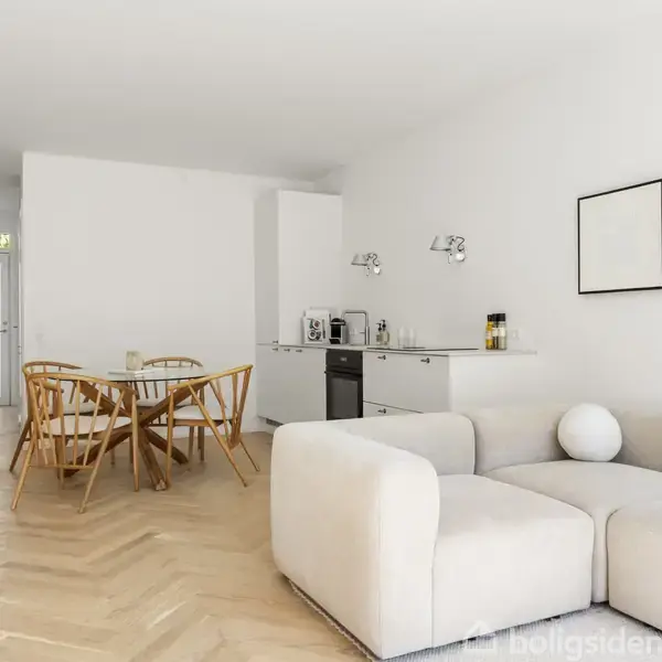 En lys stue med en beige hjørnesofa, et rundt spisebord med fire stole i træ, og et lille køkken med hvidt interiør. Væggene er dekoreret med abstrakte kunstværker.