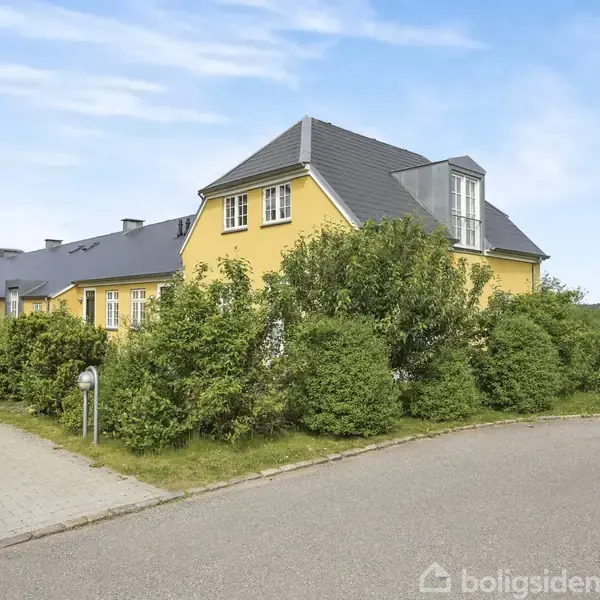 Gult rækkehus med gråt tag omkranset af frodige buske. Huset står ved en flisebelagt sti, og i baggrunden ses en grøn park og blå himmel.