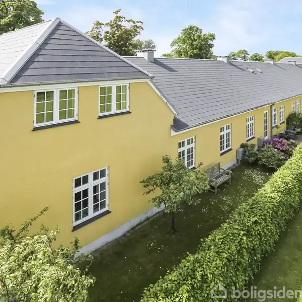 Gult rækkehus med gråt tag strækker sig langs vejen, omgivet af velplejede hække og blomsterbede, med træer og grønne områder omkring.