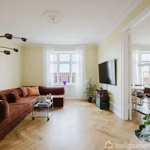 Brun sofa står i en lys stue med parketgulv; en lampe strækker sig over sofabordet. En fladskærm hænger på væggen. Der er udsigt til tilstødende spisestue gennem en døråbning.