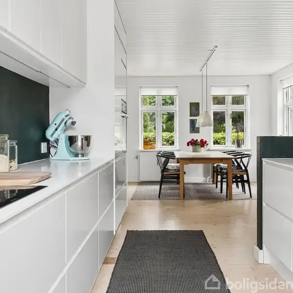 Et moderne køkken med hvide skabe har en foodprocessor ved siden af en glasbeholder med mel. Et spisebord med blomster står ved vinduet, som giver udsigt til natur.