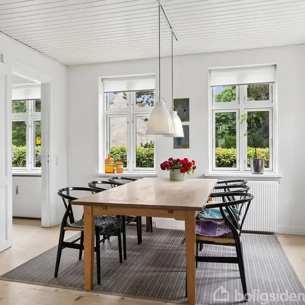 Et spisebord står i centrum med sorte stole rundt om. Bordet har en vase med blomster. Rummet er lyst med hvide vægge og store vinduer med udsigt til natur.