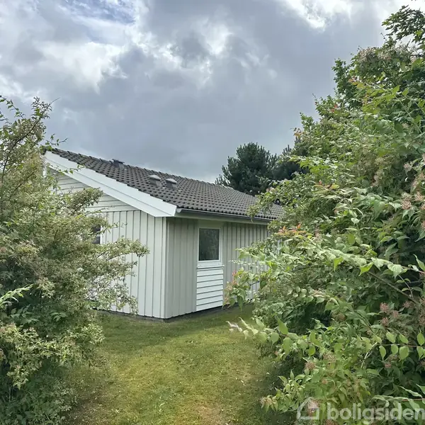 Et lille, hvidt træhus står stille omgivet af frodige buske og træer under en overskyet himmel.