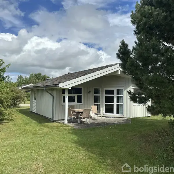 Et lille, hvidt sommerhus med terrasse og havemøbler står på en grøn græsplæne omgivet af træer og buske under en delvist skyet himmel.