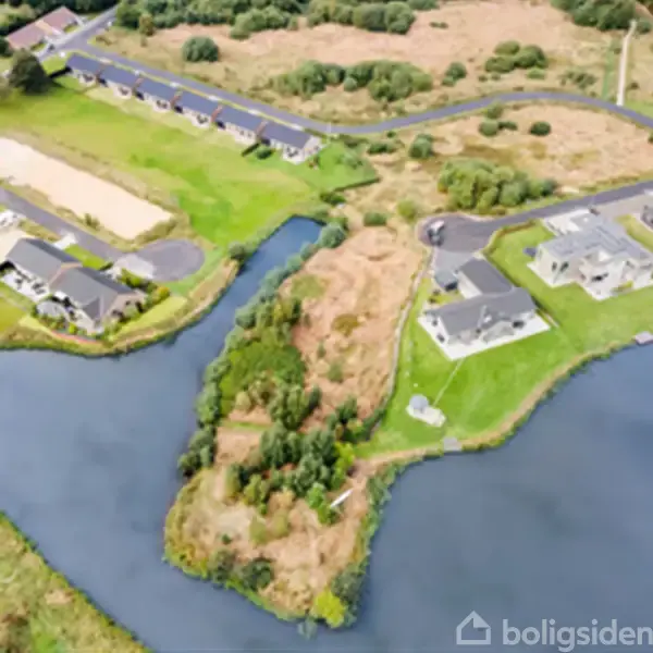 Flod omgiver et boligområde med moderne huse, placeret på frodige grønne græsarealer og små øer. Veje forbinder husene, som er omgivet af træer og åbne områder.