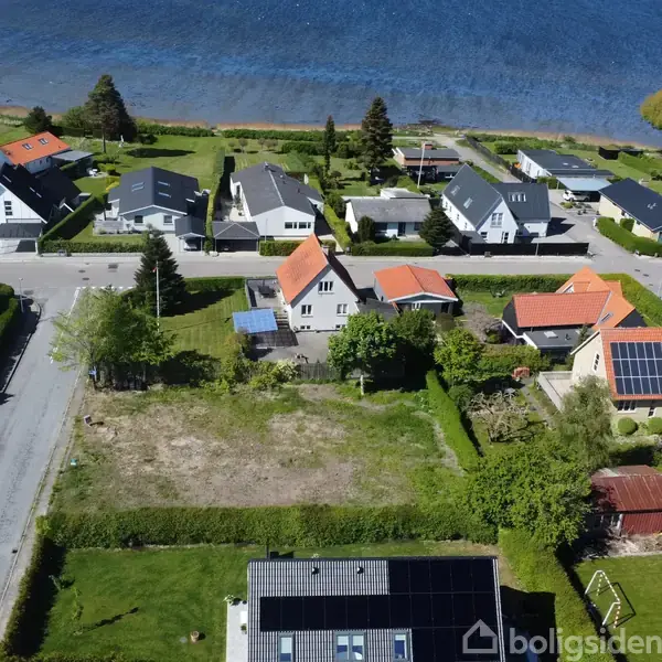 Tomt jordstykke ligger mellem to huse i et boligområde med udsigt til vand i baggrunden. Flere huse med røde og grå tage er omkring, og en vej løber langs arealet.