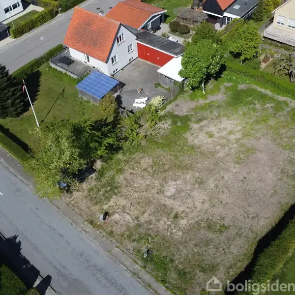 Åbent areal med spredt græs ved siden af et parcelhus med rødt tag. Stedet er omkranset af hække, og der ses en vej og flere huse i baggrunden.