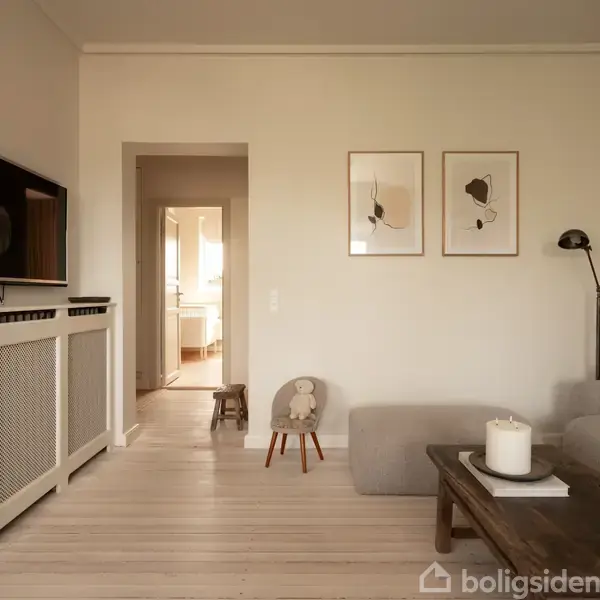 TV'en hænger på væggen, viser intet; stue med sofa og sofabord. Lys trægang leder til et køkken. To minimalistiske billeder hænger på væggen ved siden af en standerlampe.