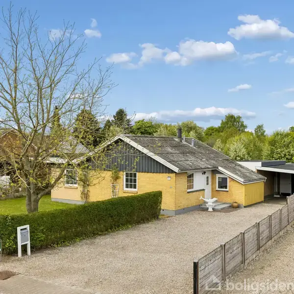 En gul murstensejendom med en carport i højre side og en gruslagt indkørsel ligger i et landområde omgivet af træer og grønne områder. En enkelt hæk markerer grænsen til fortovet.