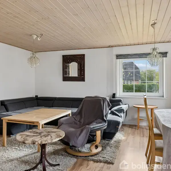 Sort hjørnesofa står i en stue med lyst trægulv og hvidt loft. Et spejl hænger på væggen. Ved siden af står et spisebord med stole og en reol ved vinduet.