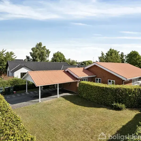 Et rødt murstenshus med rødt tegltag og dobbelt carport med tilhørende indkørsel. Det ligger i et boligkvarter omgivet af velplejede hække og grønne træer under en klar blå himmel.