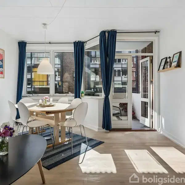 En spisestue med et bord omgivet af hvide stole. Et glas bord står i forgrunden. Store vinduer med blå gardiner giver udsigt til en bygning udenfor.