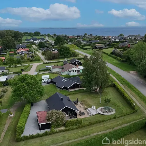 Sort hus med terrasse og have set fra luften, omgivet af grønne hække. Flere huse spredt rundt i nabolaget, tæt på kystlinjen og havudsigt i baggrunden.