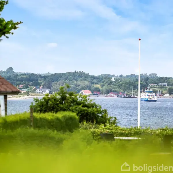 Hus tæt på vandet set fra en have. En båd sejler på en fjord i baggrunden med skovklædte bakker og spredte huse i det fjerne.