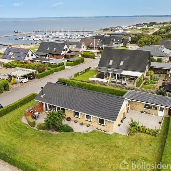 Et enkeltplanshus med sort tag ligger i en velplejet have. Huset er omgivet af andre huse med udsigt til en marina og havet i baggrunden.