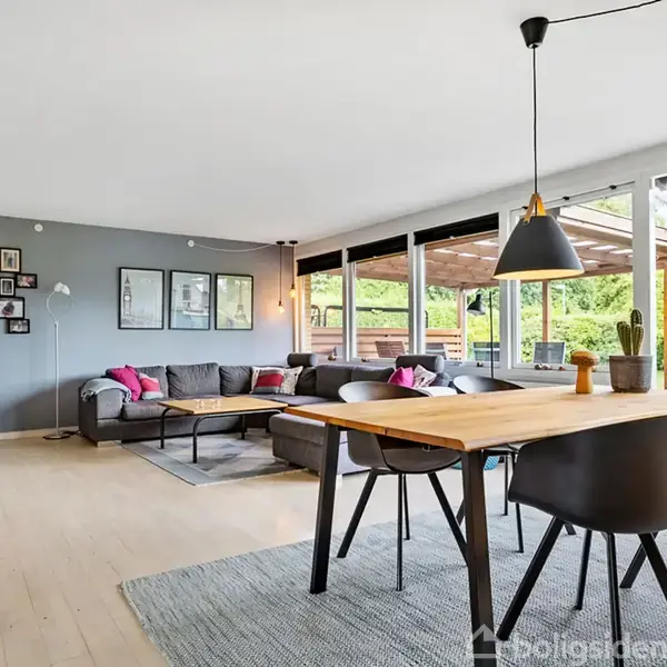 En stue med en stor grå sofa og sofabord i midten, omgivet af billeder på en grå væg. Spisebord i forgrunden, store vinduer mod en udendørs terrasse.