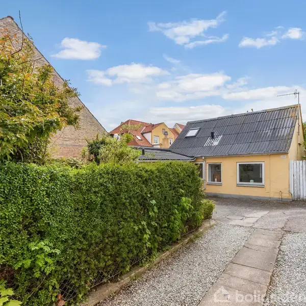 Et gult hus med sorte tagsten står stille på en grusvej. Omkranset af en grøn hæk og placeret i et villakvarter med blå himmel og få skyer.