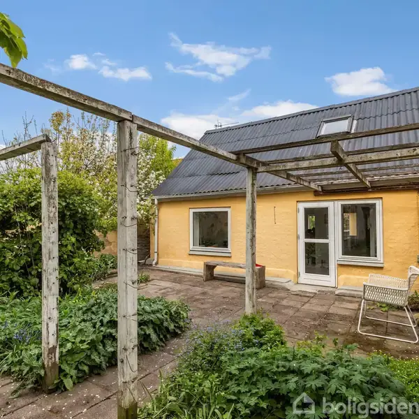 Et hus med gul facade og sort tag står i en gårdhave. Der er en pergola foran huset omgivet af grønne planter og fliser. Himmel er blå med få skyer.
