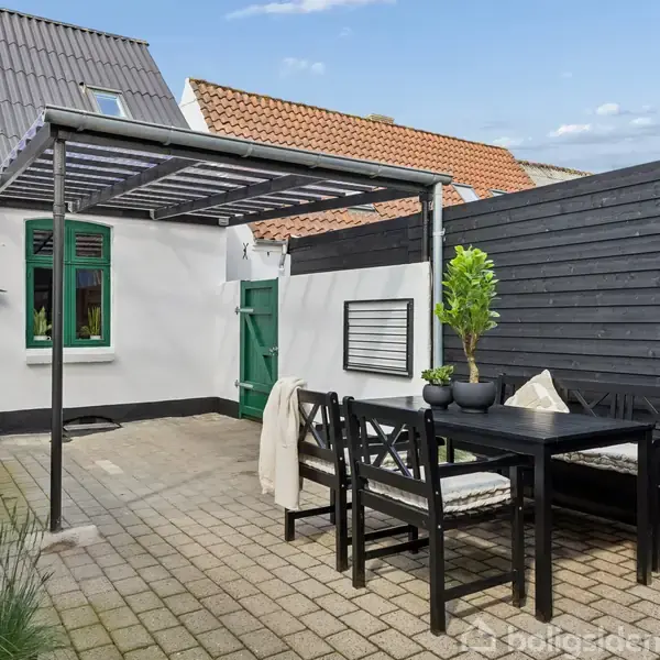 En terrasse med havebord og bænke under en pergola. Baggrunden består af hvid husmur og grå plankevæg, omkranset af forskellige grønne planter.