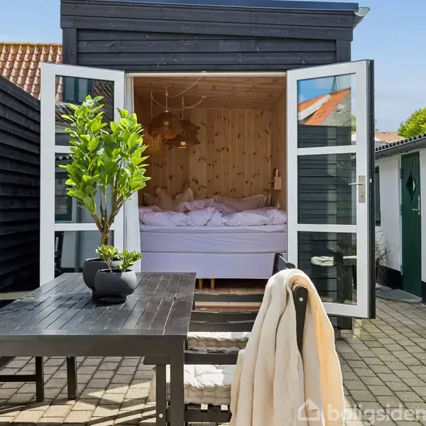 Soveværelse åbner sig til terrassen med et sort træbord og en stol. Grøn plante i potte på bordet. Husets facade er af sortmalet træ.