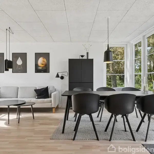 Grå sofa står i et lyst rum med åbne franske døre. Store vinduer giver udsigt til en terrasse og have. To sækkestole, rundt bord og sorte spisebordsstole er arrangeret.