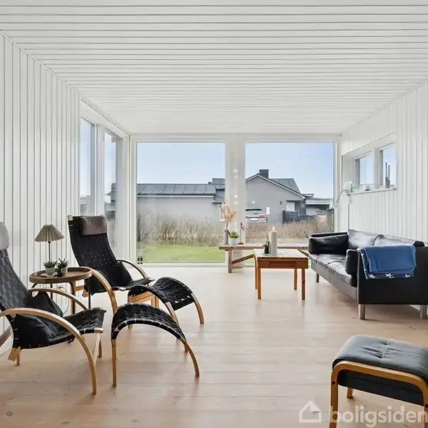 To sorte loungestole med fodskamler står i en hvid, lys stue med trævægge og store vinduer. En sort sofa og et sofabord sikrer hygge. Udsigt til have.