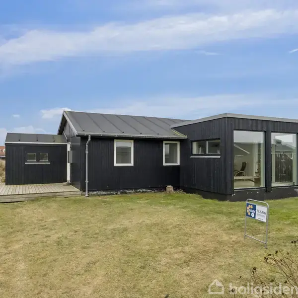 Sort træhus med store vinduer står på en grøn græsplæne med en bred terrasse. Skylinen er klar med spredte skyer. Et skilt står foran huset.