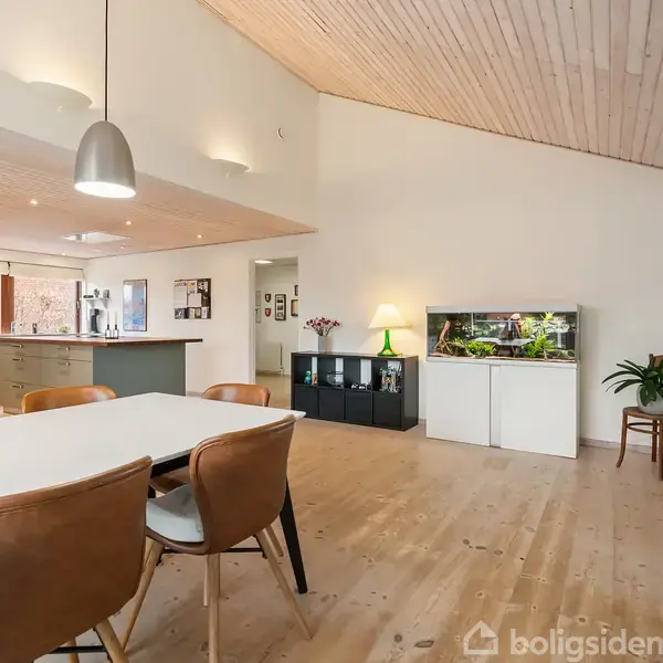Spisebord med brune stole står i en stue. En akvarium på en skænk til højre. Lyse trævægge og gulve skaber en åben og lys atmosfære. Moderne lamper hænger fra loftet.