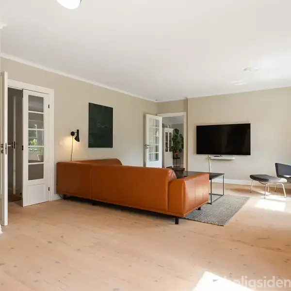En brun lædersofa står i et lyst stuehjørne med væg-tv og en kommode ved siden af. Der er et lille bord med blå blomster og moderne stole i hjørnet.