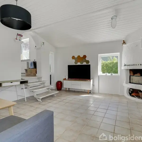 Stue med hvide vægge og loft, flisegulv. TV på hvid skænk, pejs til højre. Trægulvstrapper til venstre. Lampe hænger fra loftet. Vinduer lukker naturligt lys ind.