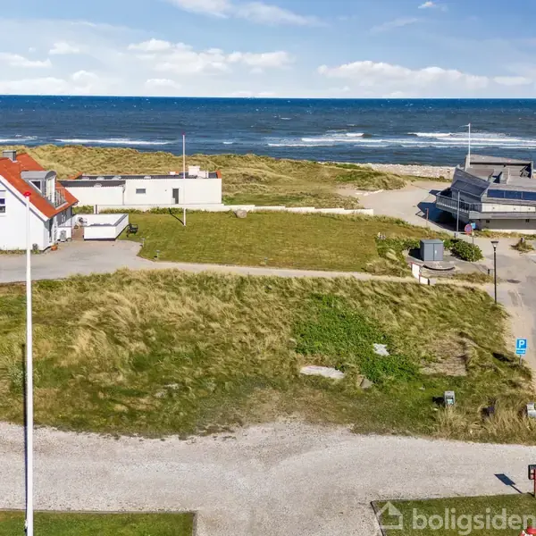 Græsbeklædte klitter ligger stille foran en række huse i en lille by. Skilt og vejlamper markerer områdets grænse. Byen strækker sig ud mod horisonten med flere bygninger.