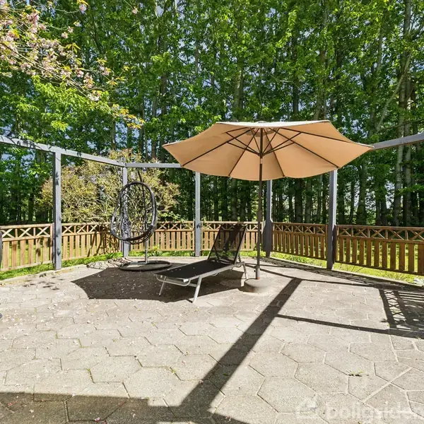 Grill dækket med overtræk står ved siden af en pergola med solstol og parasol i en flisebelagt have, omgivet af træer og hegn.