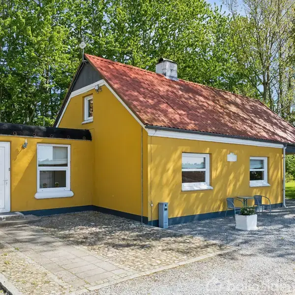 Gult hus med rødt tegltag står stille. Det er omgivet af træer og grusbelagte overflader med en lille terrasse og indgangsparti foran.