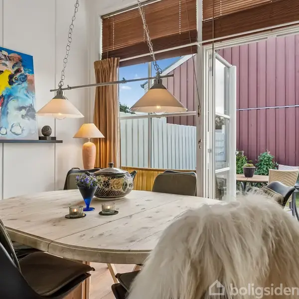 Rundt spisebord med stole omkring, dekoreret med skål og glas. På væggen hænger et farverigt abstrakt maleri. Store åbne døre fører ud til terrasse og haveredskaber.