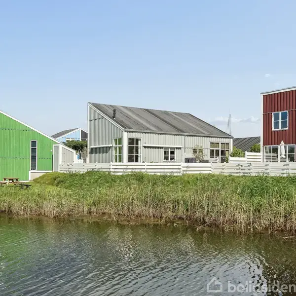 Flere farverige huse står stille langs en lille sø omgivet af grønt græs. Husene er grønne, grå og røde og har en moderne arkitektur.