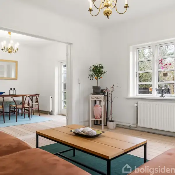 En træterrasse med et bord og fire sorte stole står ved siden af et gult hus, omgivet af en græsplæne og træer i en have.