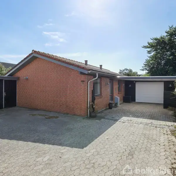 Rødt murstenshus med garage og fladt tag. Grå flisebelægning foran huset. Omgivet af grønt buskads og træer under en klar blå himmel.