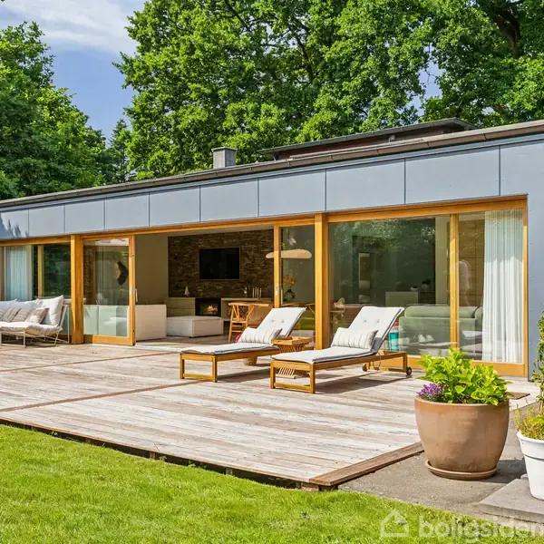 Moderne villa med store vinduespartier og træterrasse. To liggestole står på terrassen, omgivet af grøn have og store træer. Indenfor ses en stue med sofa og tv.