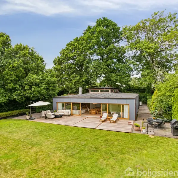 Et moderne hus omkranset af frodige træer har en stor terrasse med siddepladser og parasol. En velplejet græsplæne strækker sig foran huset, omgivet af grøn beplantning.