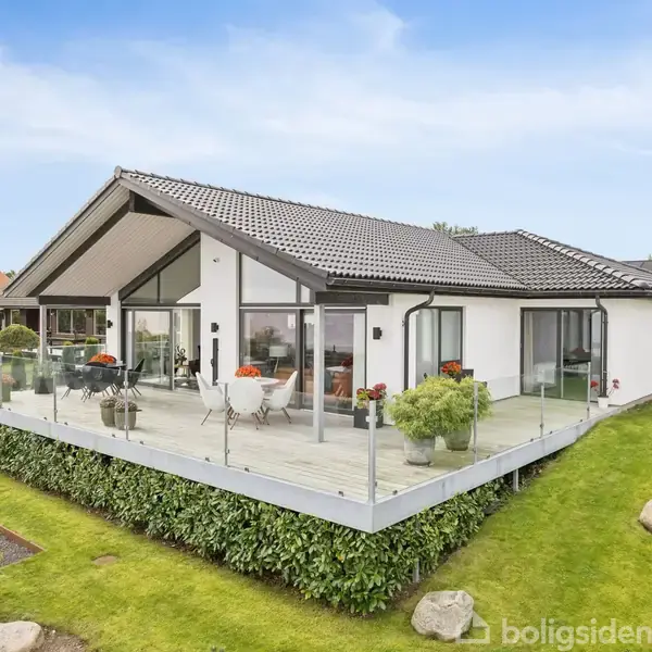 Moderne hvid villa med glasafskærmet terrasse, omgivet af velplejet græsplæne og have. Terrassen har havemøbler og planter. Huset har store vinduespartier og mørkt tag.