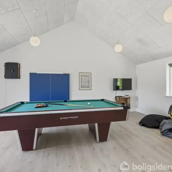 Poolbord med køer i en rummelig spillehal. En fodboldspillemaskine står ved siden af, og en fladskærm hænger på væggen. Der er et vindue med udsigt til naturen.