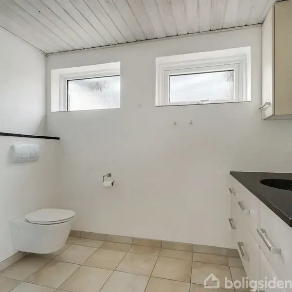 Et badeværelse med væghængt toilet, to små vinduer, en sort vask på en hvid skuffeenhed, og en bruser med matteret glasvæg. Lyse klinker dækker gulvet.