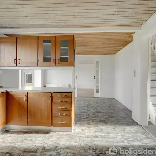 Køkkenet har træskabe og skuffer, åbner ud mod spiseområdet med et lyst flisegulv og hvidmalede vægge. Langs den ene side står en radiator, mens en trappe leder opad.