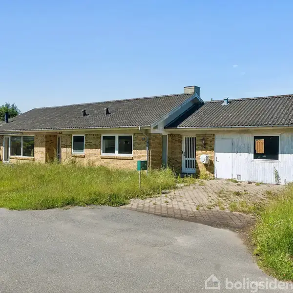 En étplanshus med brun murstensfacade og sort tag står midt i en villavej. Foran huset er der en delvist flisebelagt indkørsel med højt græs rundt omkring.